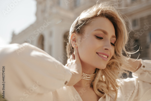 Cheerful blonde curly woman in pearl necklace and white elegant blouse smiles sincerely, looks down and poses outside. photo