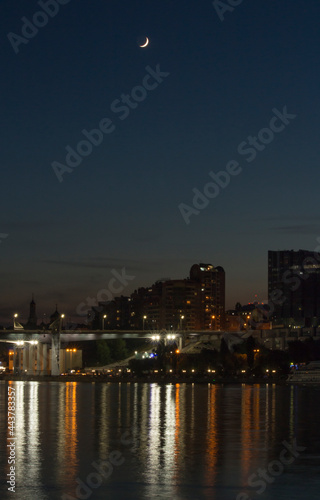 Color night photo with dark sky, young moon and cityscape. Beautiful reflection of lights in the water. Bridge and buildings at night.