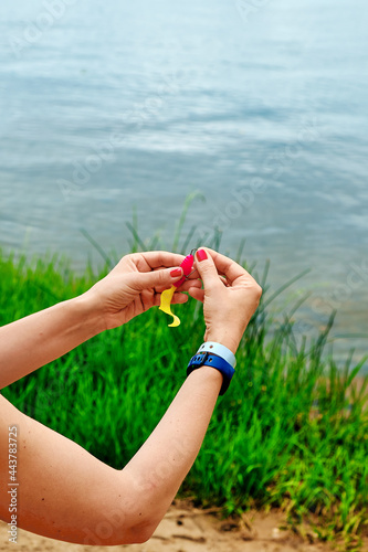female hands with red manicure putting silicone bait on a fish hook, shallow depth of field, water and coastal grass background 