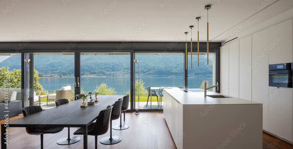 Obraz premium Interior of a modern apartment, with dining room and open kitchen. Front view. While eating you have a spectacular view of the lake. Nobody inside, the table seats six.