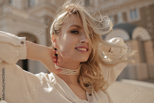 Young charming curly blonde woman in stylish white blouse and pearl necklace smiles sincerely and walks outside. photo