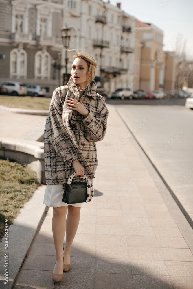 Attractive young blonde woman in checkered tweed coat and white dress walks in city center and holds handbag.