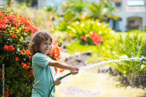 Cute little boy watering flowers in the garden at summer day. Child using garden hose. Funny kid watering plants in the yard garden.
