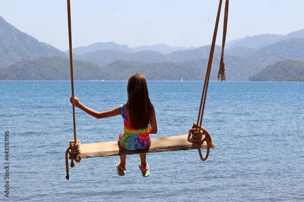 Kid girl in swimsuit swinging on a wooden swing against the blue sea and misty mountains. Beach holidays on a resort