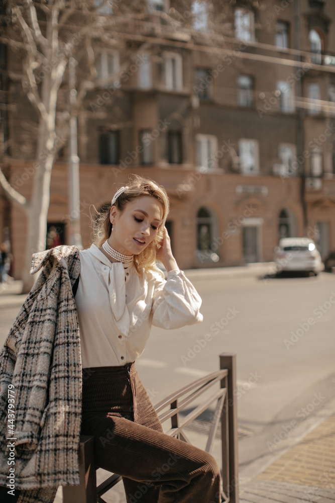 Stylish young blonde woman in brown pants, white blouse and checkered coat walk in city. Charming girl in pearl necklace poses outside.