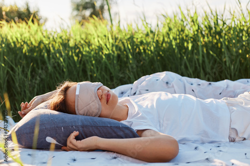Portrait of woman wearing sleeping mask and white t shirt lying on soft bed in the middle of green field or meadow, female sleeps outdoors in summertime.