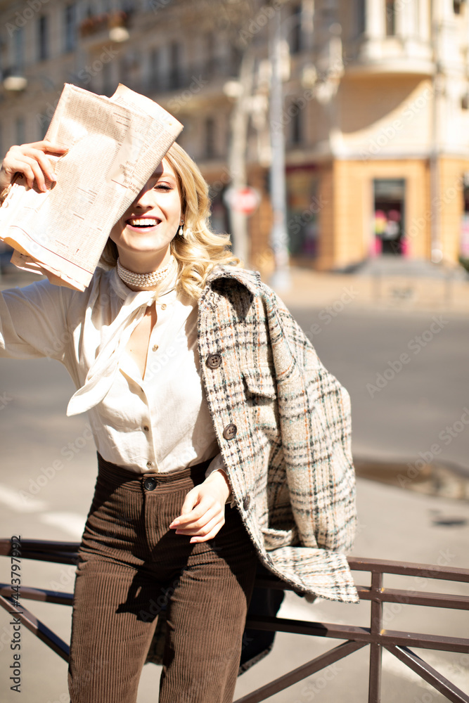Happy blonde curly woman in white blouse, brown pants and tweed checkered coat smiles and covers face using newspaper. Lady poses outside.