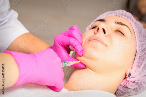 The cosmetologist makes lipolytic injection on the chin of a young woman against the double chin in a beauty salon.