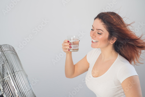 A red-haired Caucasian woman chills by the electric fan and drinks a cold drink. Climate control in the apartment