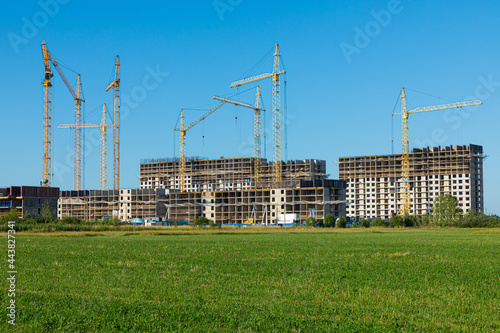 Cloudless landscape or scene of meadow with green grass and bushes with concrete modern building construction with tall yellow cranes on the background of clear blue sky