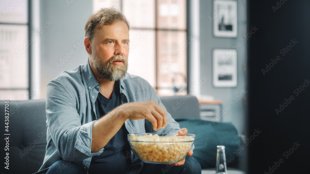 Handsome Middle Aged Bearded Man Sitting on a Couch Watches TV at Home. Focused Fan Eats Snacks, Watches TV Shows, Sports Event, Film, On Demand Streaming Service. Handsome Middle Aged Bearded Man Sitting on a Couch Watches TV at Home. Focused Fan Eats Snacks, Watches TV Shows, Sports Event, Film, On Demand Streaming Service.