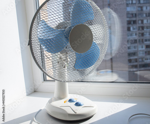 a white fan stands on the window in the apartment during the day. Cooling system, room air conditioning, hot summer 2021. Window fan, shortage