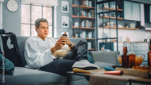 Photography Handsome Man Sitting on a Couch Watches Ice Hockey Game on TV, uses Smartphone App for Score, Bet, Statistics, Eats Snacks, Watching Championship