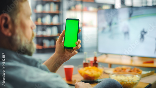 At Home Ice Hockey Fan Holds Green Screen Chroma Key Smartphone, Friends Watch Game on TV, Cheer for Favourtite Sports Team to Win Championship. Professional Club Play. Over the Shoulder Close-up photo