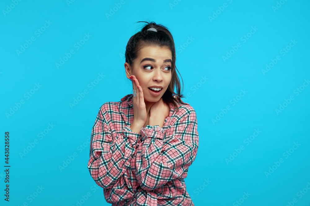 Fototapeta premium Beautiful wondered woman keeps hand on cheek looks surprisingly aside dressed in trendy clothes reacts on shocking news isolated over blue background