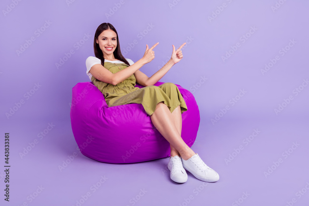 Full body photo of cheerful young happy woman point finger empty space sit bean bag isolated on purple color background Full body photo of cheerful young happy woman point finger empty space sit bean bag isolated on purple color background