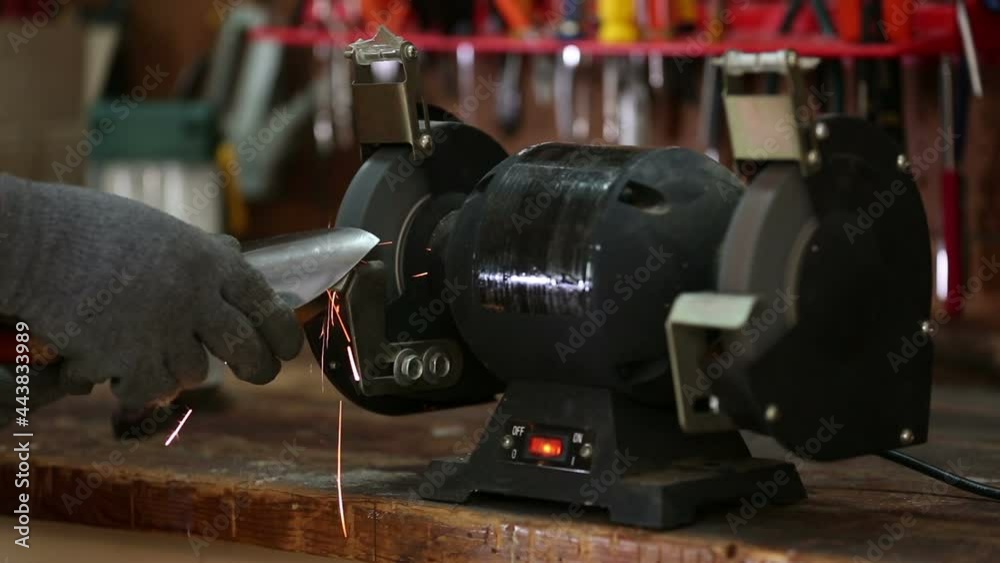 Sharpening a spade hand trowel wearing gloves on a bench grinder with sparks flying on workbench