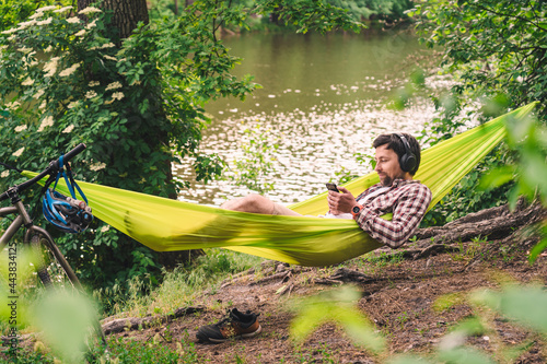 Man on bicycle trip at camping by lake is relaxing in green hammock while listening to music. Active recreation theme in nature. Hipster cyclist with headphones having fun in hammock by river