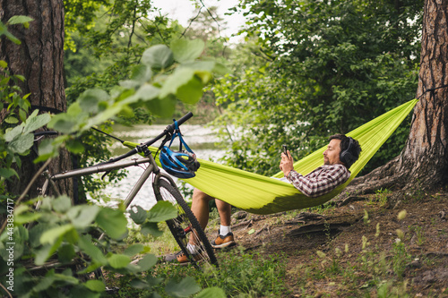 Young caucasian man resting in hammock, listening to music on headphones and using smartphone afterwards on bicycle in forest near the lake. Audio healing. Idyllic place. Travel, camping in nature