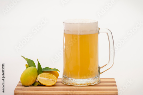 Jarra de cerveza con refresco de limón y limones con hojas, sobre una tabla de madera y fondo blanco.