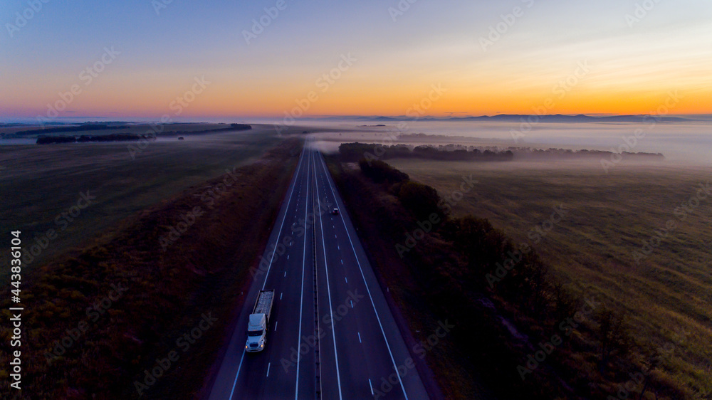 Fototapeta premium View from above. An asphalt road is located among endless green meadows. Automobile road in the fog against the background of dawn.