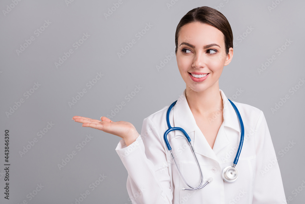 Photo of cute positive young doc woman dressed white coat smiling looking holding arm empty space isolated grey color background Photo of cute positive young doc woman dressed white coat smiling looking holding arm empty space isolated grey color background