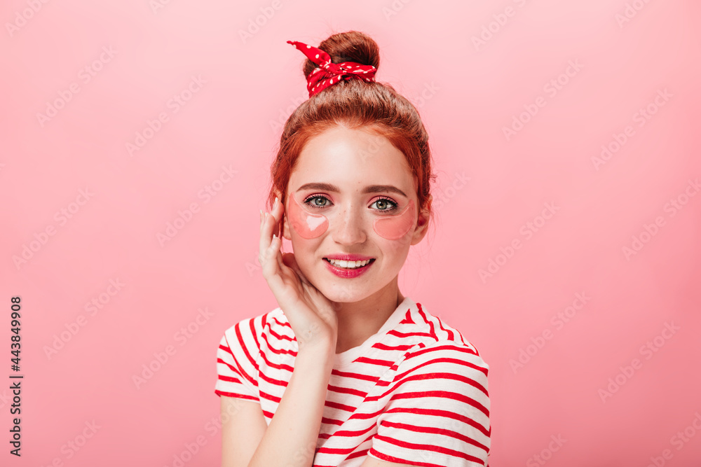 Trendy caucasian girl with eye patches looking at camera with smile. Studio shot of ginger woman in striped t-shirt isolated on pink background. Trendy caucasian girl with eye patches looking at camera with smile. Studio shot of ginger woman in striped t-shirt isolated on pink background.