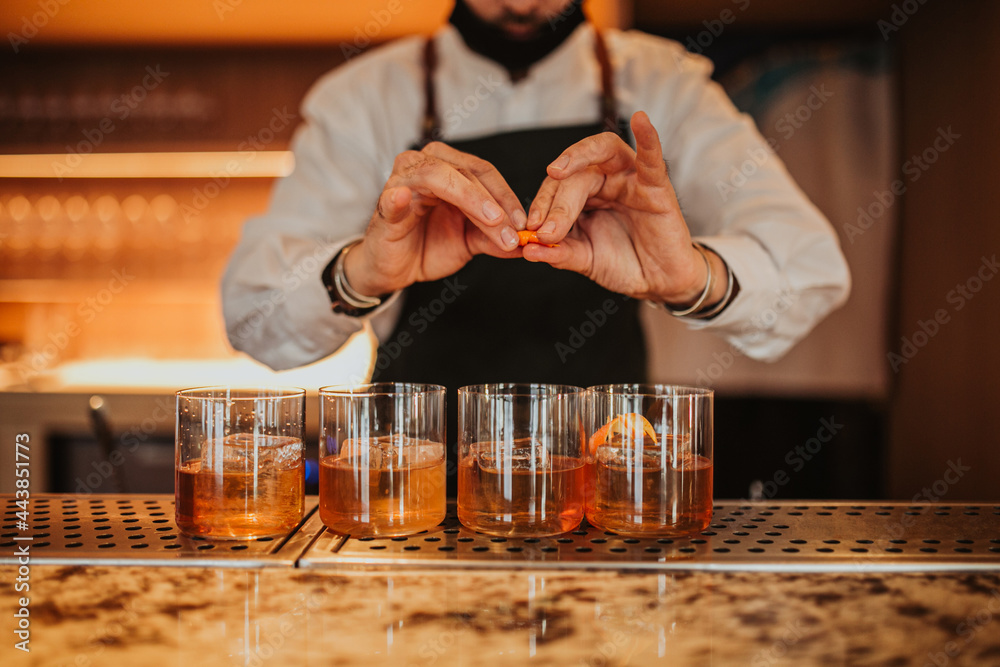 A close up shot of a bartender in a green apron making Old Fashioned cocktails in a restaurant. Focus on glasses and hands. Concept of hospitality and bartending. Horizontal image. A close up shot of a bartender in a green apron making Old Fashioned cocktails in a restaurant. Focus on glasses and hands. Concept of hospitality and bartending. Horizontal image.