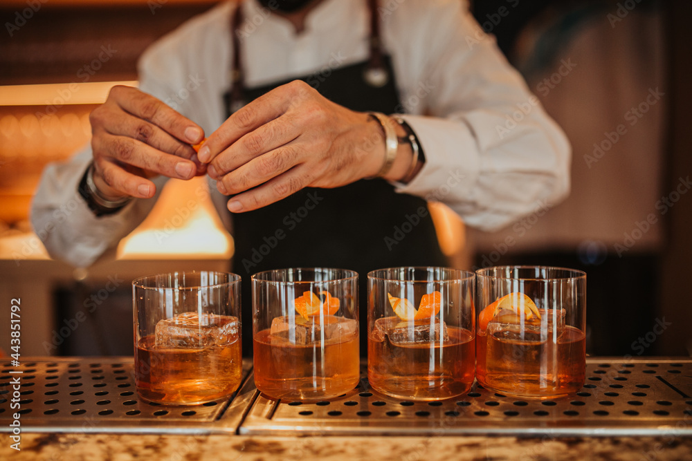 A close up shot of a bartender in a green apron making Old Fashioned cocktails in a restaurant. Focus on glasses and hands. Concept of hospitality and bartending. Horizontal image.