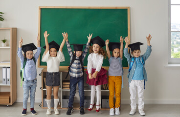 Kids graduate. Portrait of funny little kindergarten or junior high school graduates who are dressed in mortar boards. Kids having fun in the classroom on the background of the school board.