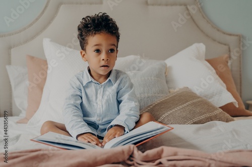 Cute curly haired afro american little boy sitting on bed with book