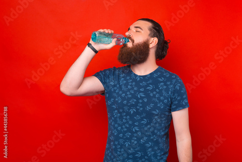 Portrait of young bearded man drinking water from bottle standing on red background.