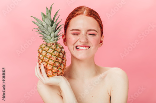 Front view of glad woman with pineapple posing with closed eyes. Studio shot of excited ginger girl holding fruit on pink background.