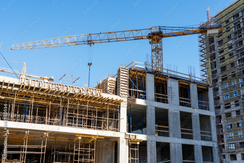Construction cranes and unfinished residential buildings against clear blue sky. Housing construction, apartment block with scaffolding