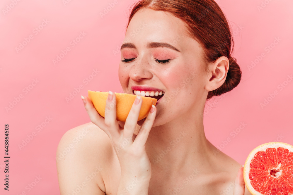 Pleased ginger girl eating grapefruit. Studio shot of sensual woman enjoying fruits on pink background. Pleased ginger girl eating grapefruit. Studio shot of sensual woman enjoying fruits on pink background.