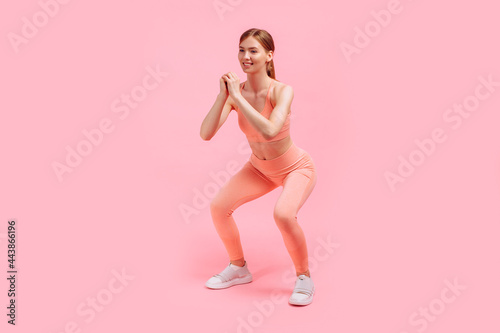 young athletic woman in leggings and top squats on a pink background, sports, healthy lifestyle.