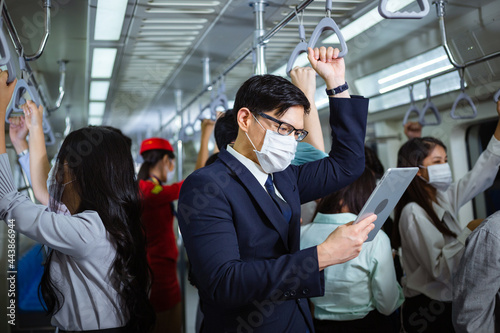 Asian man wearing medical face mask reading news or checking work on computer tablet in subway with crowded people. new normal lifestyle during coronavirus pandemic