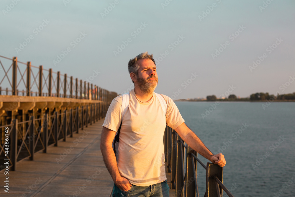 Fototapeta premium A young man is standing on the pier against the backdrop of the seascape and looking at the sunset. Huelva, Spain.