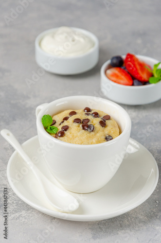 Mug cake with chocolate chips, fresh berries and cream on a gray concrete background. Copy space.