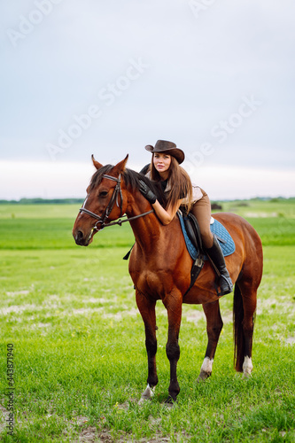 Young woman dressed in riding clothes and hat riding brown horse in green field