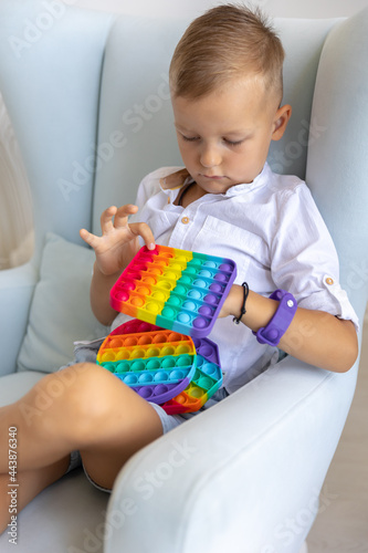 Portrait blonde boy with pop it sensory toy different forms. Boy presses colorful rainbow squishy soft silicone bubbles in children's playroom. Stress and anxiety relief. Trendy fidgeting game.
