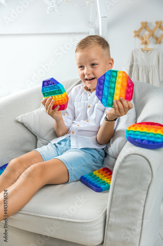 Portrait blonde boy with pop it sensory toy different forms. Boy presses colorful rainbow squishy soft silicone bubbles in children's playroom. Stress and anxiety relief. Trendy fidgeting game.