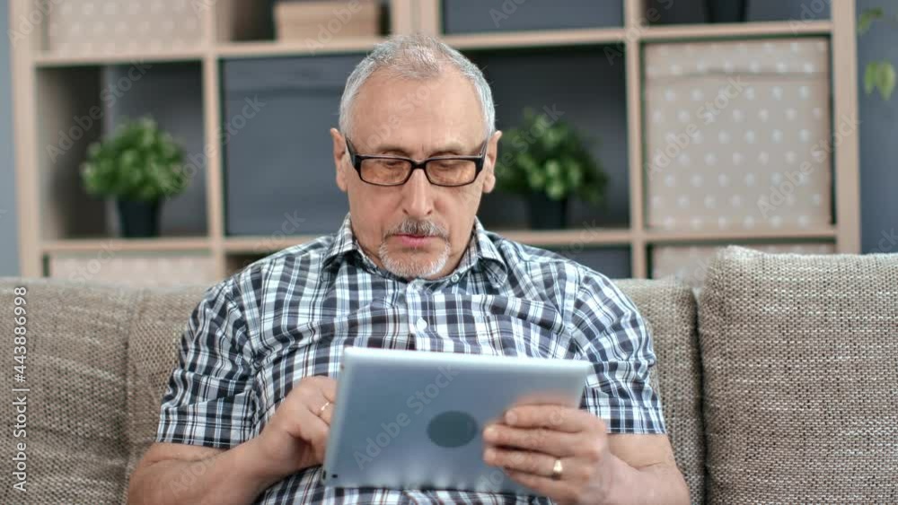 Smiling elderly 70s man studying digital tablet computer pad playing games reading e book on sofa Smiling elderly 70s man studying digital tablet computer pad playing games reading e book on sofa