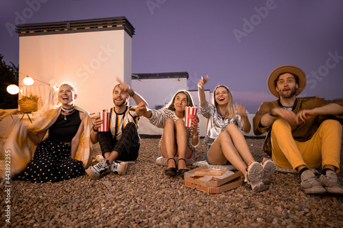 Young people watching football match or some film with popcorn outdoors on a rooftop terrace at night. Open air cinema concept