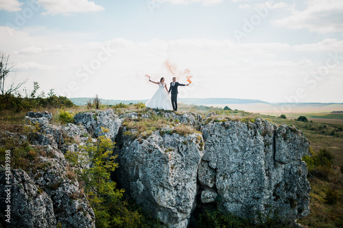 Newlyweds stand against the background of rocks and hold colorful smoke in their hands