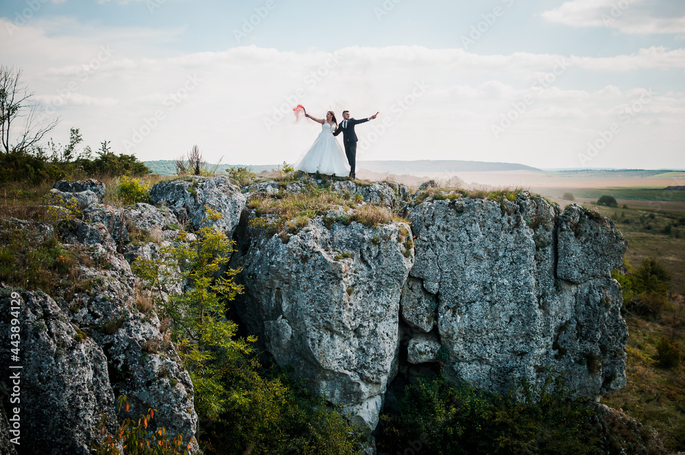 Fototapeta premium Newlyweds stand against the background of rocks and hold colorful smoke in their hands