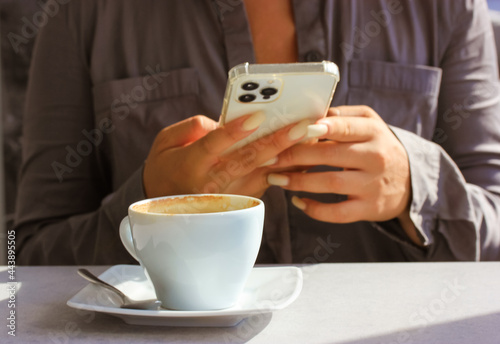 A young modern businesswoman in cafe is holding a white Iphone 12 Pro smartphone, typing a text message, surfing for information in the Internet. A cup of coffee, table.