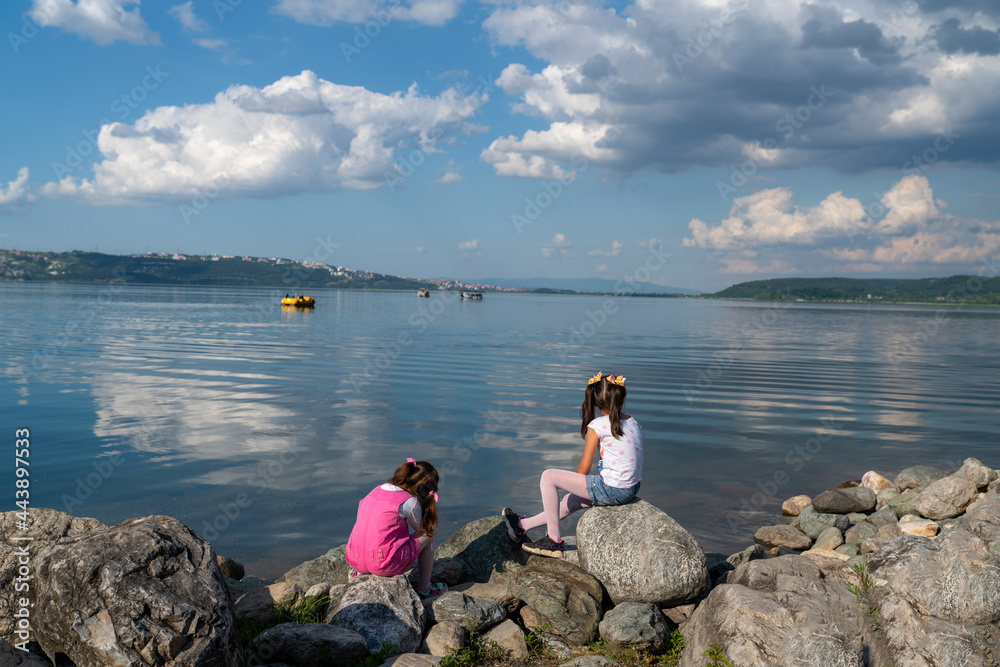 Fototapeta premium Back view of two girls sitting on the shore.