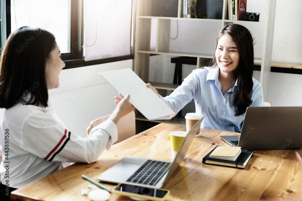 Two female company employees happily smiled at each other as they handed each other folders. Two female company employees happily smiled at each other as they handed each other folders.