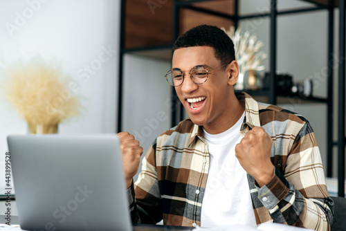 Amazed excited latino male freelancer or office worker, sitting at his work desk at office, using laptop, shouts and rejoice in success, win or good profit, looking at laptop and gesturing hands